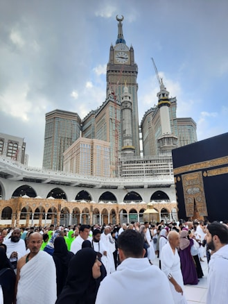 A large crowd of people dressed in traditional attire gather around a significant religious site with a tall clock tower and impressive architectural structures in the background. The scene is bustling with activity and a sense of reverence.