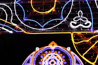 Intricate neon lights arranged in artistic patterns with vivid blue, orange, and white lines creating geometric and abstract shapes against a dark background.