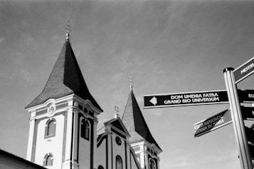 A black-and-white photograph featuring two church towers with pointed spires. In the foreground, there is a street sign with multiple directional arrows pointing towards cultural and religious sites.
