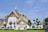 A grand and ornate temple with intricate architectural details including gold and red accents against white walls. The temple features a tiered roof with spires reaching toward the sky. There are manicured gardens and topiary trees in the foreground, enhancing the serene and majestic atmosphere.