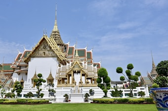 A grand and ornate temple with intricate architectural details including gold and red accents against white walls. The temple features a tiered roof with spires reaching toward the sky. There are manicured gardens and topiary trees in the foreground, enhancing the serene and majestic atmosphere.