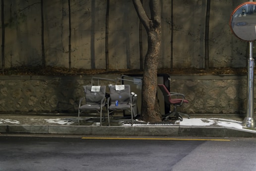 Several chairs are placed on a sidewalk next to a tree, with a stone wall and metal mirror nearby. There are two chairs that appear to be stacked with pieces of paper on them, and an upright office chair. Snow covers parts of the ground, and the area is dimly lit, suggesting a cold or late time of day.