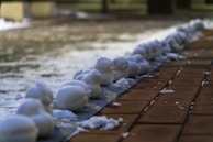 A row of small snow sculptures arranged neatly along the edge of a paved walkway. The sculptures appear to be shaped into various forms, possibly resembling animals or abstract shapes. The ground around the sculptures is wet, suggesting melting snow, and there are small patches of snow scattered around.