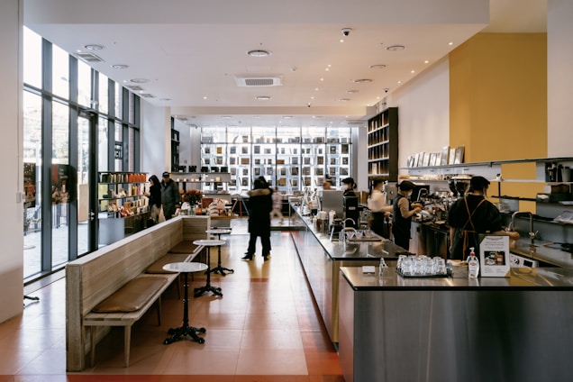 A modern cafe with high ceilings and large windows letting in natural light. The space is open and features high shelves filled with books and various items. Several people are present, with some working behind the counter preparing drinks. The seating area has simple wooden benches and small tables.