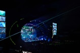 A concert stage is illuminated with vibrant lights and screens displaying various images. The atmosphere is dynamic, with beams of light cutting across the venue filled with a large audience. The stage is equipped with multiple screens and a circular arrangement of lights, creating an immersive visual experience.