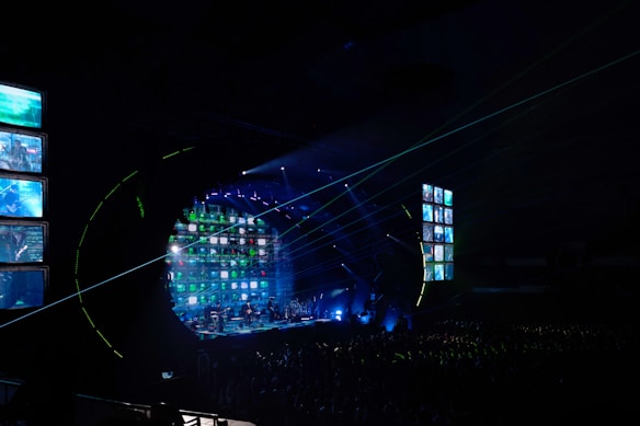 A concert stage is illuminated with vibrant lights and screens displaying various images. The atmosphere is dynamic, with beams of light cutting across the venue filled with a large audience. The stage is equipped with multiple screens and a circular arrangement of lights, creating an immersive visual experience.