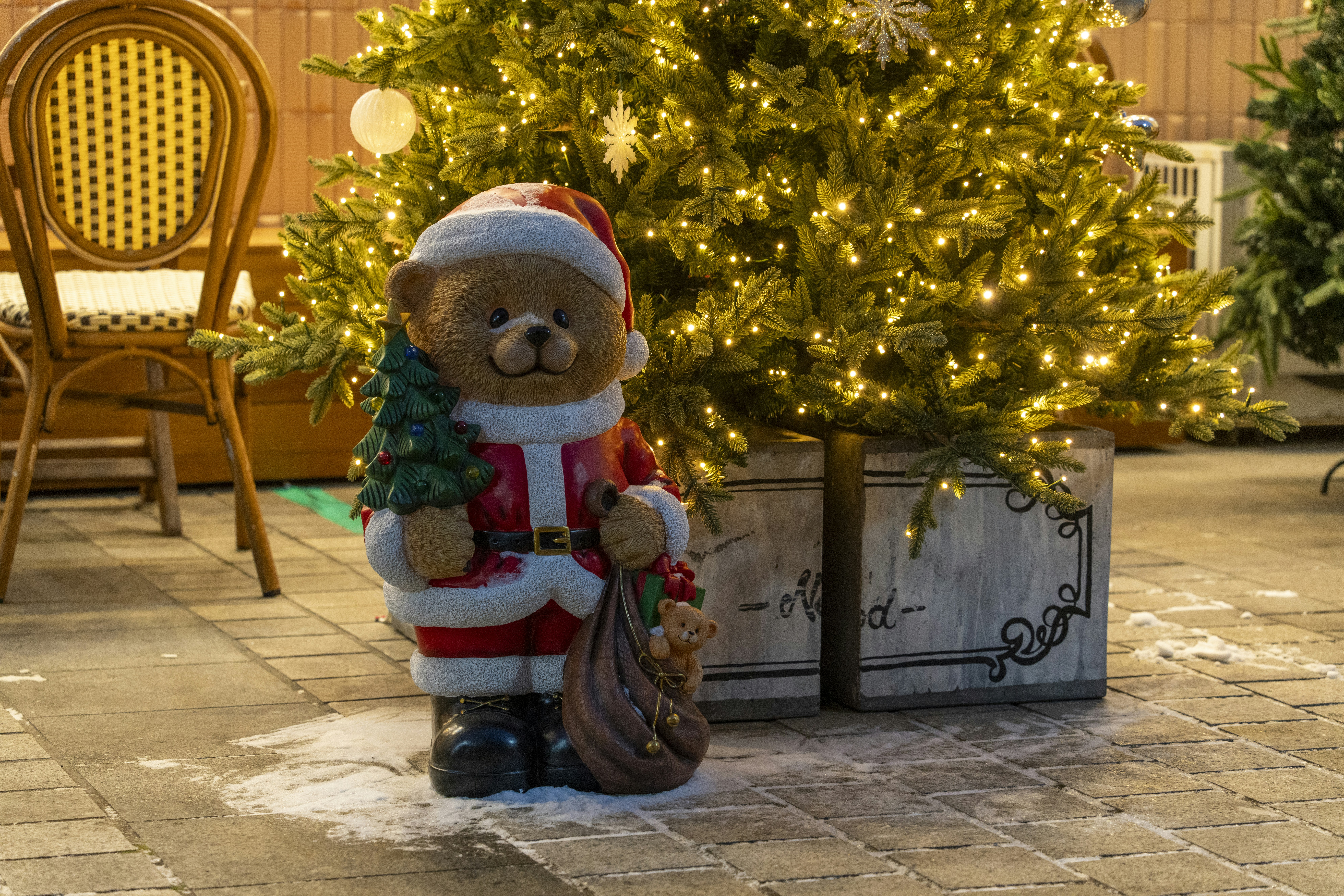  festive Christmas scene featuring a teddy bear statue dressed as Santa Claus, holding a small Christmas tree and a sack with a toy bear inside. The bear is placed in front of a decorated and lit Christmas tree, with warm white lights.