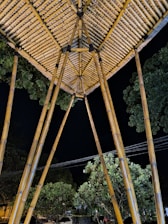A bamboo structure with poles supporting a geometric, thatched roof. Surrounding the structure are lush green trees against a dark night sky.