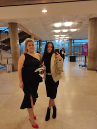 Two women dressed in elegant black dresses pose in a well-lit interior space with modern architecture. One woman is wearing red high heels and holding papers, while the other is taking a selfie with a smartphone. The background shows geometric ceiling lights, large windows, and a staircase.