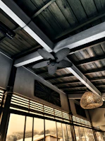 An interior scene with a wooden ceiling featuring a decorative ceiling fan and a wicker light fixture. Large floor-to-ceiling windows reveal an outdoor view with trees and a building, all in a warm, muted color palette.