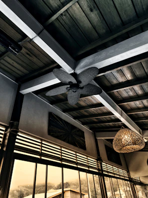 An interior scene with a wooden ceiling featuring a decorative ceiling fan and a wicker light fixture. Large floor-to-ceiling windows reveal an outdoor view with trees and a building, all in a warm, muted color palette.