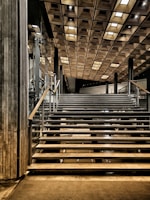 A modern staircase leading upwards, featuring sleek metal railings and steps. The ceiling above is adorned with a wooden grid pattern, creating a geometric aesthetic. Glass walls and polished surfaces contribute to the contemporary design, with strategic lighting adding depth and contrast.