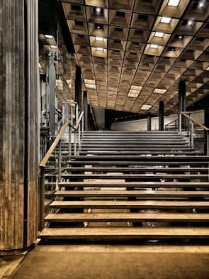 A modern staircase leading upwards, featuring sleek metal railings and steps. The ceiling above is adorned with a wooden grid pattern, creating a geometric aesthetic. Glass walls and polished surfaces contribute to the contemporary design, with strategic lighting adding depth and contrast.
