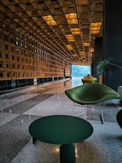 A modern and elegant interior space featuring a long corridor with a geometric wooden ceiling and walls. The corridor is lined with large floor-to-ceiling windows on one side, allowing natural light to stream in. In the foreground, there are stylish green chairs and a matching table on a polished stone floor. There are a few indoor plants adding a touch of nature to the contemporary design.