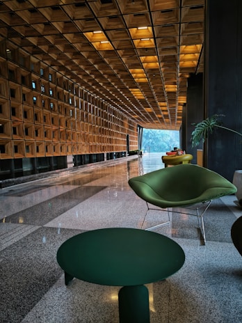 A modern and elegant interior space featuring a long corridor with a geometric wooden ceiling and walls. The corridor is lined with large floor-to-ceiling windows on one side, allowing natural light to stream in. In the foreground, there are stylish green chairs and a matching table on a polished stone floor. There are a few indoor plants adding a touch of nature to the contemporary design.