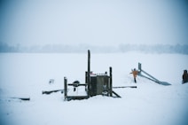 A snowy landscape with a playground covered in snow. The scene includes a wooden play structure and a slide. Two children play nearby, bundled in winter clothing with one in an orange coat. The white snow dominates the landscape, giving a serene and cold ambiance.