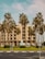 A modern multi-story hotel building with large windows is surrounded by tall palm trees. The hotel has a beige facade and a prominent sign that reads 'Hotel'. The area around the building contains well-maintained greenery and a few small structures. The sky is partly cloudy, and a busy road with traffic signs runs in front of the hotel.