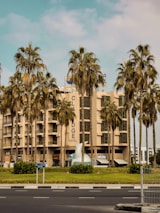 A modern multi-story hotel building with large windows is surrounded by tall palm trees. The hotel has a beige facade and a prominent sign that reads 'Hotel'. The area around the building contains well-maintained greenery and a few small structures. The sky is partly cloudy, and a busy road with traffic signs runs in front of the hotel.