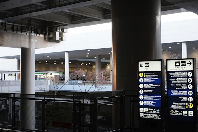 An urban setting featuring a bus stop with directional signage and list of bus routes. The area is covered, supported by large columns and features various signs, including digital displays. The background includes other parts of the infrastructure visible through openings.
