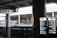 An urban setting featuring a bus stop with directional signage and list of bus routes. The area is covered, supported by large columns and features various signs, including digital displays. The background includes other parts of the infrastructure visible through openings.