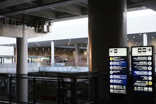 An urban setting featuring a bus stop with directional signage and list of bus routes. The area is covered, supported by large columns and features various signs, including digital displays. The background includes other parts of the infrastructure visible through openings.