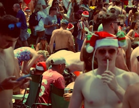 A crowded outdoor gathering of predominantly shirtless people, wearing festive Santa hats. Many are standing or sitting in folding chairs, engaging in various activities, some holding drinks and wearing sunglasses.