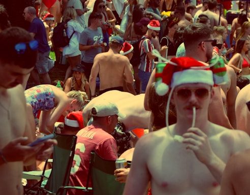 A crowded outdoor gathering of predominantly shirtless people, wearing festive Santa hats. Many are standing or sitting in folding chairs, engaging in various activities, some holding drinks and wearing sunglasses.