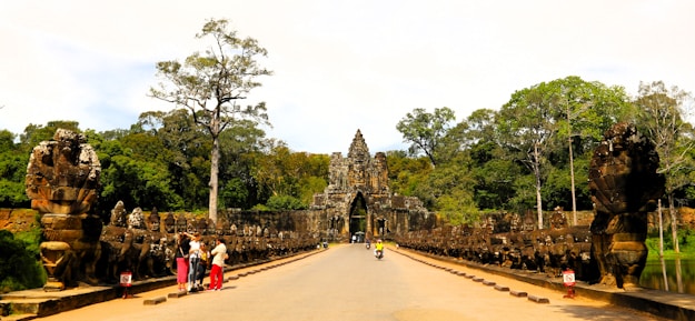 An ancient, intricately carved stone gate towers over a broad walkway flanked by large statues and lush green trees in the background. Several people can be seen walking or taking pictures, while some wear colored clothing that stands out against the earthy tones of the structure.