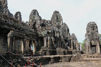 Ancient stone structures with intricate carvings and towering faces create a mysterious and intriguing atmosphere. A person is walking along the stone path, which is flanked by red ropes.