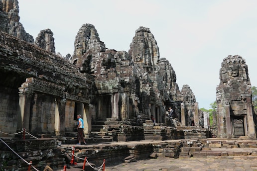 Ancient stone structures with intricate carvings and towering faces create a mysterious and intriguing atmosphere. A person is walking along the stone path, which is flanked by red ropes.