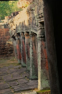 Ancient stone columns with intricate carvings line a weathered walkway. The structure is aged, covered with moss, and partially illuminated by natural light filtering through the surrounding foliage.