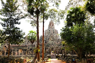 A majestic ancient stone temple adorned with intricate carvings surrounded by lush green trees. The architecture reflects a grand, historical aesthetic amid a sunlit jungle setting.