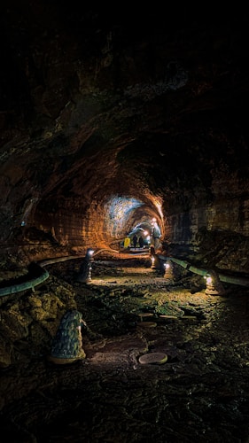 A dimly lit underground tunnel with rugged, rocky walls and floor. The tunnel extends into the distance with an arched ceiling illuminated by small, warm lights. There are a few people in the distance, and the tunnel is decorated with what appear to be small sculptures or stone formations.