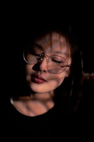 A person is partially illuminated by dappled shadows, with a serene expression and wearing large round glasses. The focus is on the face with closed eyes, creating a sense of calm and introspection. The background is dark, enhancing the play of light and shadow.