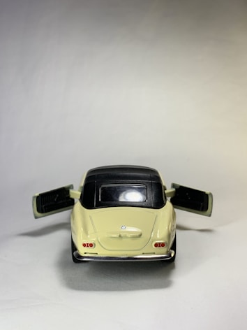 A model car is positioned against a neutral backdrop, with its doors open. The car has a cream-colored body and a black roof, and it's designed to resemble a classic automobile.