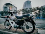 A motorcycle is parked on a brick pavement by the side of an urban street. In the background, there are modern buildings and cars are visible on the road. Trees line the street behind the motorcycle, creating an urban yet slightly natural setting.
