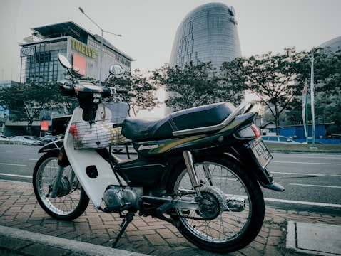 A motorcycle is parked on a brick pavement by the side of an urban street. In the background, there are modern buildings and cars are visible on the road. Trees line the street behind the motorcycle, creating an urban yet slightly natural setting.
