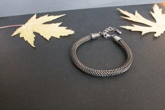 A woven metallic bracelet with a clasp is placed on a dark background. Two dried, pale yellow maple leaves are laid symmetrically on each side of the bracelet, enhancing the contrast between the elements.