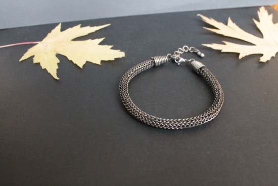 A woven metallic bracelet with a clasp is placed on a dark background. Two dried, pale yellow maple leaves are laid symmetrically on each side of the bracelet, enhancing the contrast between the elements.