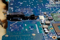 A close-up view of a circuit board with various electronic components such as capacitors, resistors, and connectors. The surface is covered in intricate patterns of pathways and solder points, with a metallic sheen and areas of green and blue.