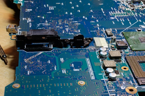 A close-up view of a circuit board with various electronic components such as capacitors, resistors, and connectors. The surface is covered in intricate patterns of pathways and solder points, with a metallic sheen and areas of green and blue.