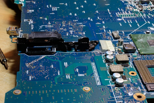 A close-up view of a circuit board with various electronic components such as capacitors, resistors, and connectors. The surface is covered in intricate patterns of pathways and solder points, with a metallic sheen and areas of green and blue.
