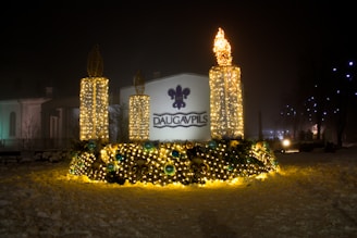 A nighttime scene features a decorative outdoor display with three large, vertical, candle-like structures adorned with twinkling yellow lights. The base is lavishly decorated with greenery and additional lights, accented by spherical ornaments in shades of blue. The word 'Daugavpils' is prominently displayed on a backdrop suggesting a town or city name.