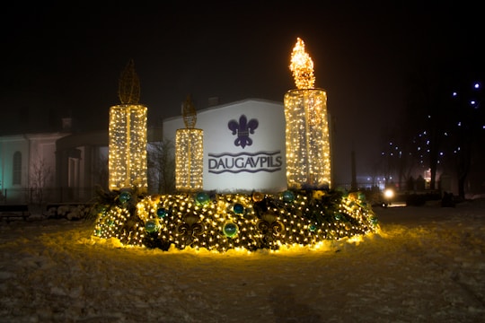 A nighttime scene features a decorative outdoor display with three large, vertical, candle-like structures adorned with twinkling yellow lights. The base is lavishly decorated with greenery and additional lights, accented by spherical ornaments in shades of blue. The word 'Daugavpils' is prominently displayed on a backdrop suggesting a town or city name.