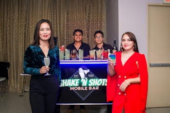 Two women holding colorful drinks stand in front of a mobile bar setup. Behind them, two bartenders are smiling, while various cocktails are displayed on the bar. The setting appears to be indoors with a curtain backdrop.
