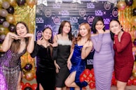 A group of six women dressed in colorful party dresses are standing in front of a balloon-covered backdrop. The women are posing playfully, making peace signs and other gestures. The background is decorated with a mix of gold, black, and shiny, iridescent balloons, along with branded logos.