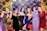 A group of six women dressed in colorful party dresses are standing in front of a balloon-covered backdrop. The women are posing playfully, making peace signs and other gestures. The background is decorated with a mix of gold, black, and shiny, iridescent balloons, along with branded logos.