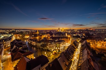 A vibrant cityscape captured at twilight, showcasing a mix of historical and modern buildings illuminated by warm golden lights. The sky features a gradient from deep blue to orange as the sun sets on the horizon. A festive atmosphere is suggested by the presence of illuminated trees lining the streets, indicating possible holiday decorations.