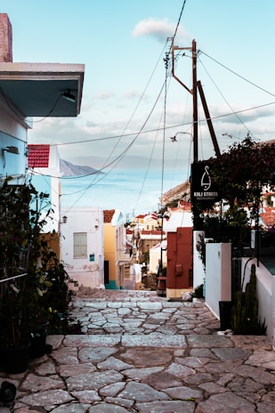 A narrow cobblestone street descends toward the sea, lined with colorful buildings featuring white, red, and pastel facades. Overhead, power lines stretch across and a clear blue sky meets the horizon. A sign reading 'Kali Strata Restaurant' is visible, with various potted plants and a cactus adding greenery.