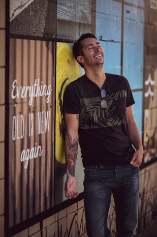 A person with a tattoo on their arm is leaning casually against a wall with a colorful mural. They are wearing a black t-shirt, jeans, and sunglasses hanging from the shirt. The person is smiling, suggesting a relaxed and happy mood. The wall features a large text saying 'Everything old is new again.'